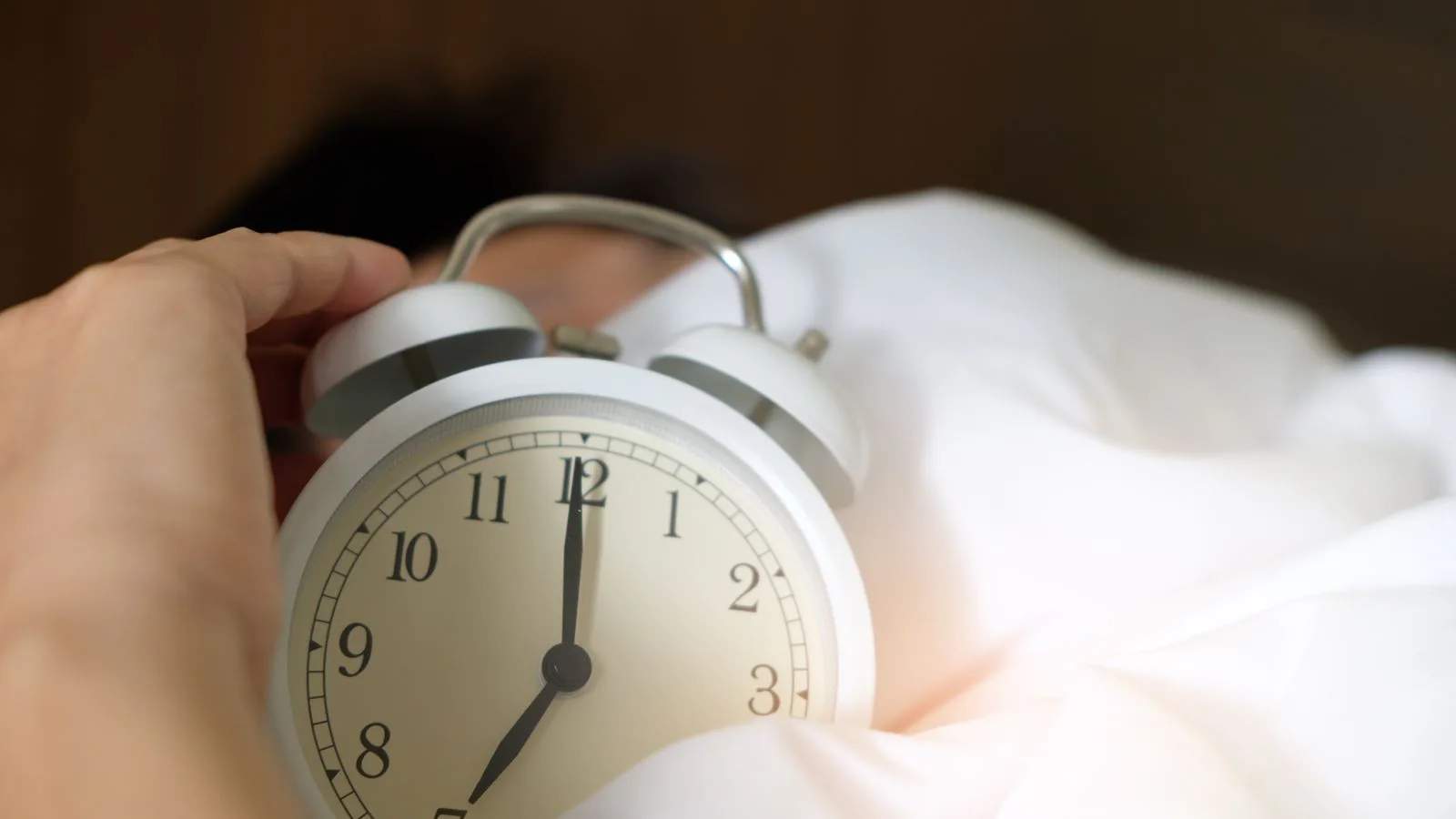 Uma mão estende-se para um despertador a tocar, sobre a cama.
