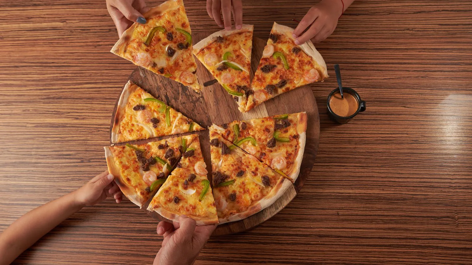 Várias pessoas tiram fatias de uma pizza partilhada numa mesa de madeira.