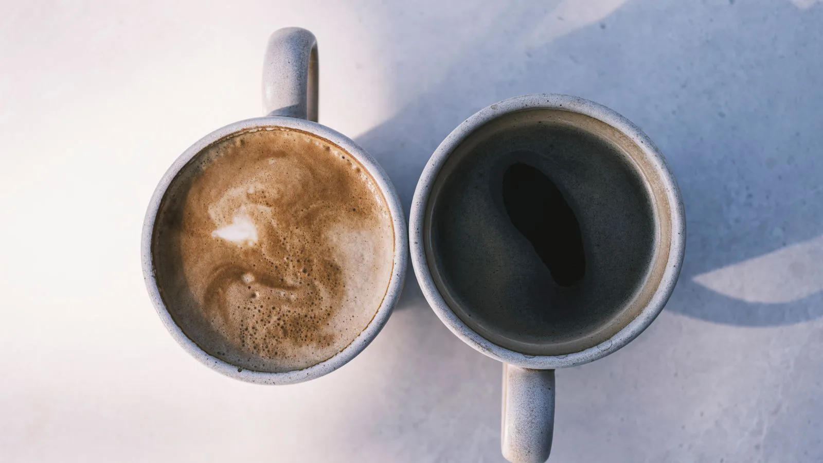 Two mugs of coffee on a sunlit table, seen from above.