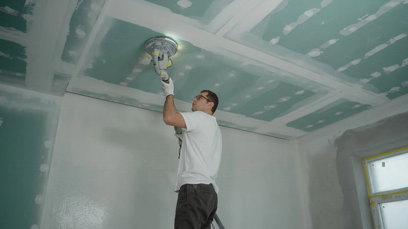 A worker smoothing a plasterboard ceiling indoors.