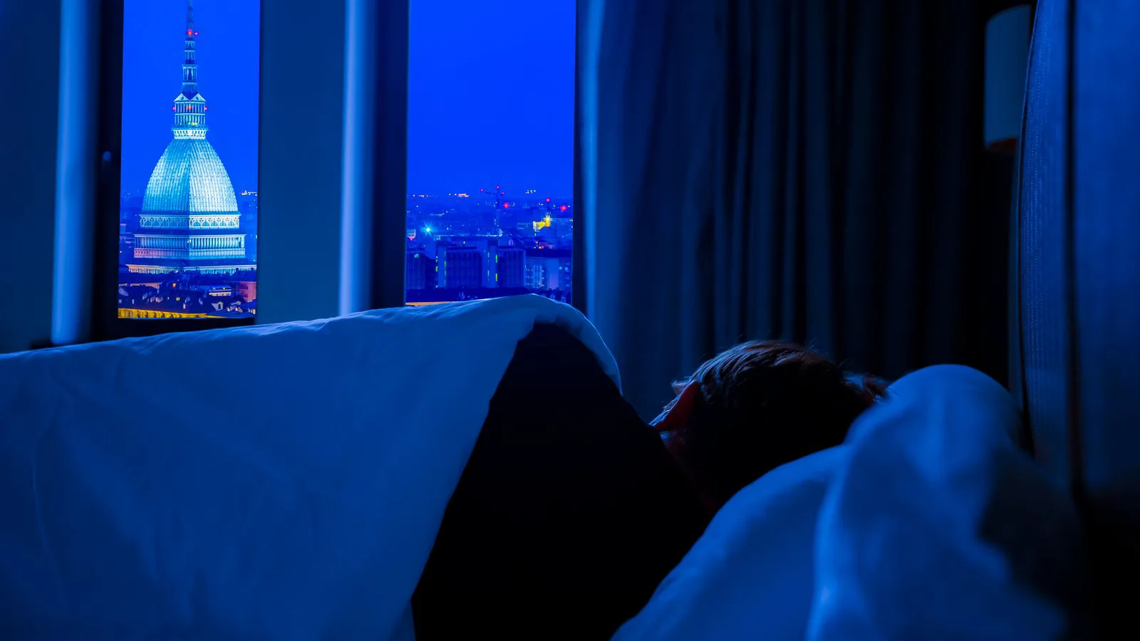 A darkened bedroom with a city skyline glowing through the window.