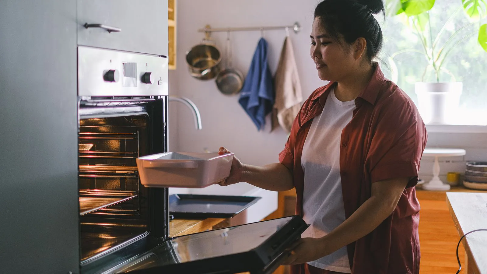 A woman sliding a dish into an oven in a sunlit kitchen.