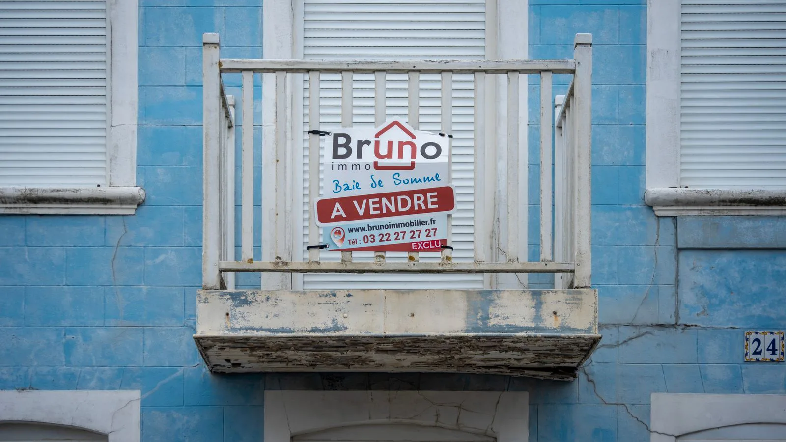 A worn blue facade with an estate agent's board on the balcony.