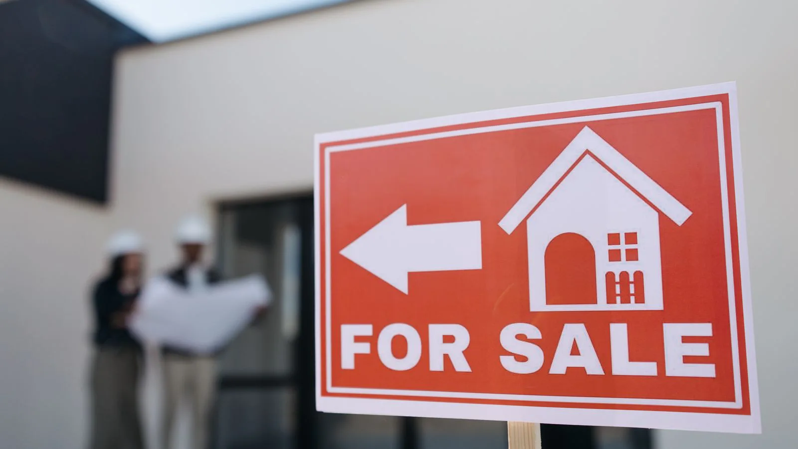 A for-sale board in front of a house, two people talking behind it.