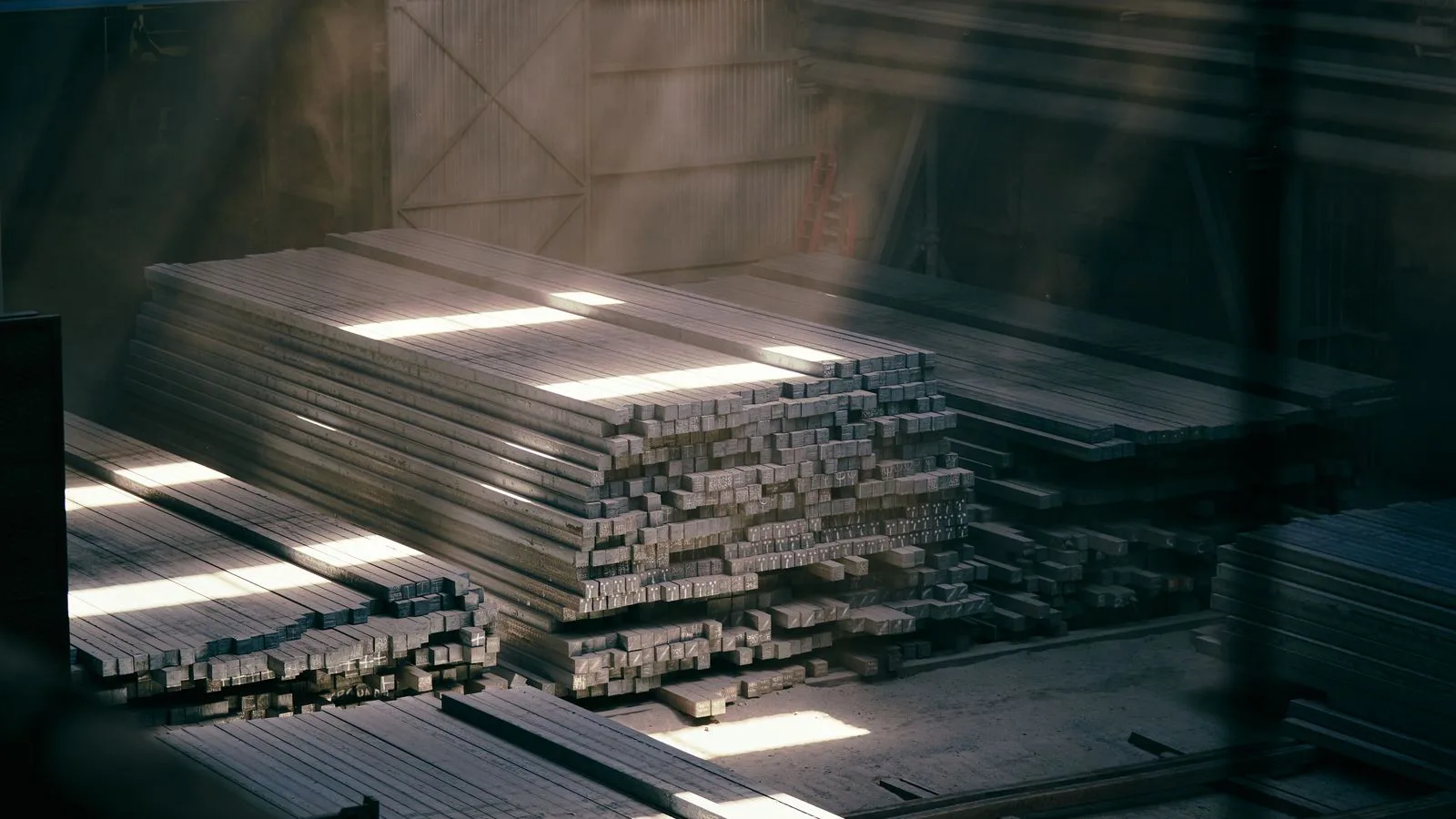 Stacked steel sections in a warehouse, lit from a high window.