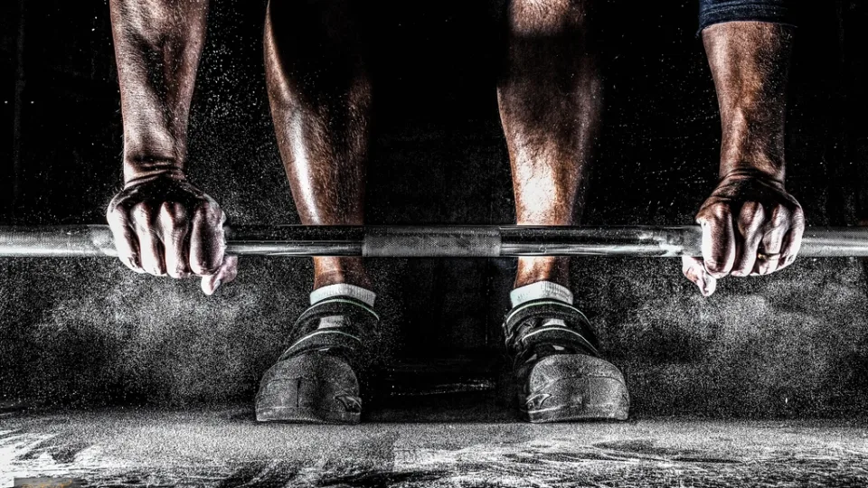Two hands gripping a barbell on the floor in a cloud of chalk dust.