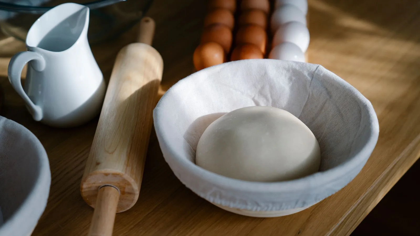 A bowl of dough beside a rolling pin, a jug and eggs on a wooden table.